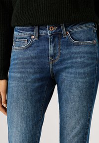 Sinised teksad kitsas lõikes, klassikalise viie tasku disainiga, vasksete detailidega ja kerge kulumise efektiga tekstuuri lisamiseks. Kandetud musta džempriga.