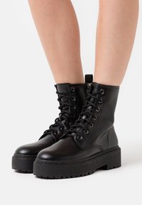Mustat nahka ankle bootsit, joissa on nauhakiinnitys edessä, hopeiset silmukat ja platform-pohjat. Sileä pinta ja ommeltu detaljointi sivuilla.