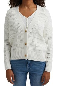 Vit stickad cardigan med ett texturerat mönster, med V-ringning, knappstängning och ribbat nederkant. Buren över en vit topp och jeans.
