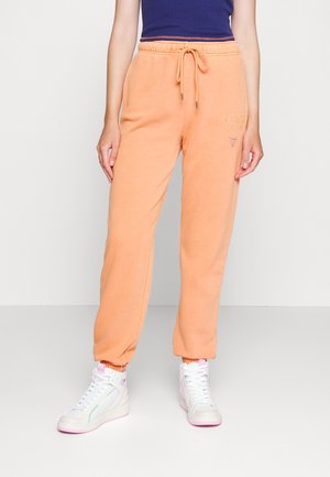 Persoon draagt perzikkleurige joggerbroek met trekkoord in de taille, een marineblauw top en witte hoge sneakers, staand op een witte vloer.