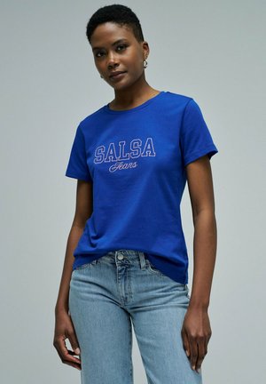 Mujer con el pelo corto, que lleva pendientes de aro plateados, una camiseta azul "SALSA Jeans" y vaqueros azul claro, de pie frente a un fondo gris liso.