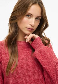 Punane kudum sweater tekstuuriga pinnaga, ümmarguse kaelusega ja pikkade varrukatega, millel on soonikkoes mansetid. Mudel poseerib käsi lõua juures.