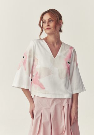 Žena s červenými vlasmi oblečená v bielej blúzke s výstrihom do V a ružovými kvetinovými motívmi a plisovanú svetloružovú sukňu, stojaca pred jednoliatym pozadím.