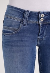 Istutetut siniset farkut, joissa on hieman kulunut pesu, kaksi etutaskua, kaksoisnappisuljin ja kontrastiväriset tikkaukset.