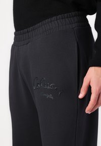 Dark grey sweatpants with an elastic waistband, featuring embroidered branding on the side and a smooth texture.
