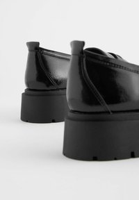 Scarpe nere in pelle verniciata con suola nera spessa e leggermente piatta, caratterizzate da una texture liscia e dettagli di cucitura minimi.
