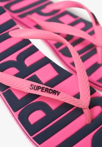 Roze slippers met marineblauwe strepen, met een geprofileerde voetbed en rubberen banden. "SUPERDRY" is in reliëf op een band gedrukt.