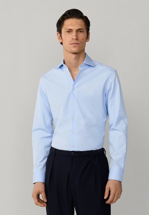 Man die een lichtblauw overhemd met lange mouwen draagt, ingestopt in een zwarte gerimpelde broek, staand tegen een effen grijze achtergrond.