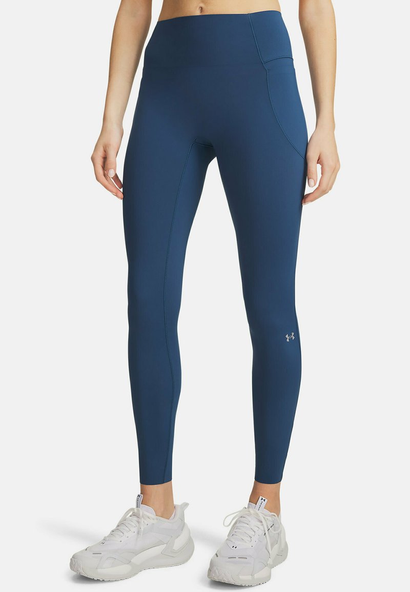 Femme portant des leggings de sport bleu marine taille haute avec une poche latérale et des chaussures de course blanches, debout devant un fond blanc.