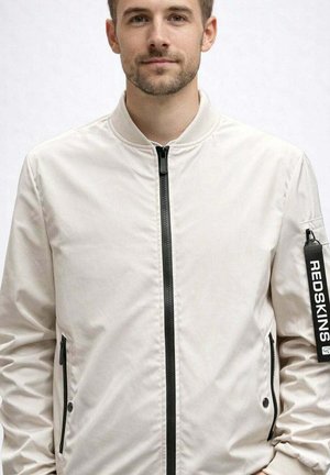 Homme portant une veste zippée beige clair avec une fermeture éclair et des poches noires, une étiquette "REDSKINS" noire et blanche sur la manche gauche.