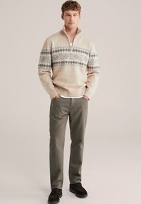 Beżowy sweter z półzamkiem, zdobiony niebieskim i czarnym wzorem. Połączony z oliwkowymi spodniami i ciemnymi butami.