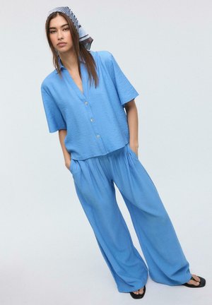 Kobieta stojąca z rękami w kieszeniach, ubrana w luźną niebieską koszulę na guziki, pasujące szerokie spodnie, pasiasty chustę na głowę i czarne sandały.