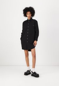 Abito camicia nero con maniche lunghe, bottone anteriore e tasca sul petto. Indossato con scarpe platform nere e calzini bianchi. Tessuto liscio.