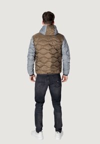 Pufferjakke med en brun quiltet krop og lysegrå ærmer, med hætte og ribbede manchetter, parret med mørke jeans og hvide sneakers.