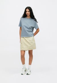 Jasnoniebieski bawełniany T-shirt z krótkimi rękawami, w połączeniu z kremową spódnicą dżinsową. Model nosi białe i zielone sneakersy z szarymi skarpetkami.