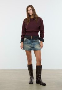 Giovane donna che indossa un maglione bordeaux, una minigonna di jeans e stivali alti fino al ginocchio in pelle marrone scuro, in piedi contro uno sfondo chiaro e uniforme.