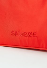 Rød nylonpose med det pregete logoet "SAMSØE" i ton-i-ton tråd, med en glatt, matt tekstur.