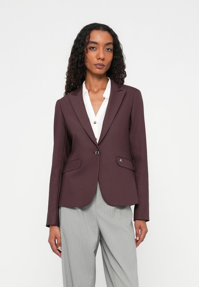 Blazer bordeaux con design su misura, due tasche frontali e chiusura a un bottone. Indossato sopra una camicia bianca, dalla texture morbida.