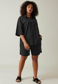 Camisa negra de lino de manga corta con botones, combinada con pantalones cortos negros. La tela parece ligera y con textura. Usada con sandalias negras.