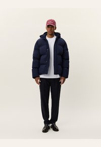 Marineblaue Steppjacke mit einem gesteppeten Design, Kapuze und vorderen Knöpfen, kombiniert mit einem weißen T-Shirt, dunklen Hosen und schwarzen Schuhen.