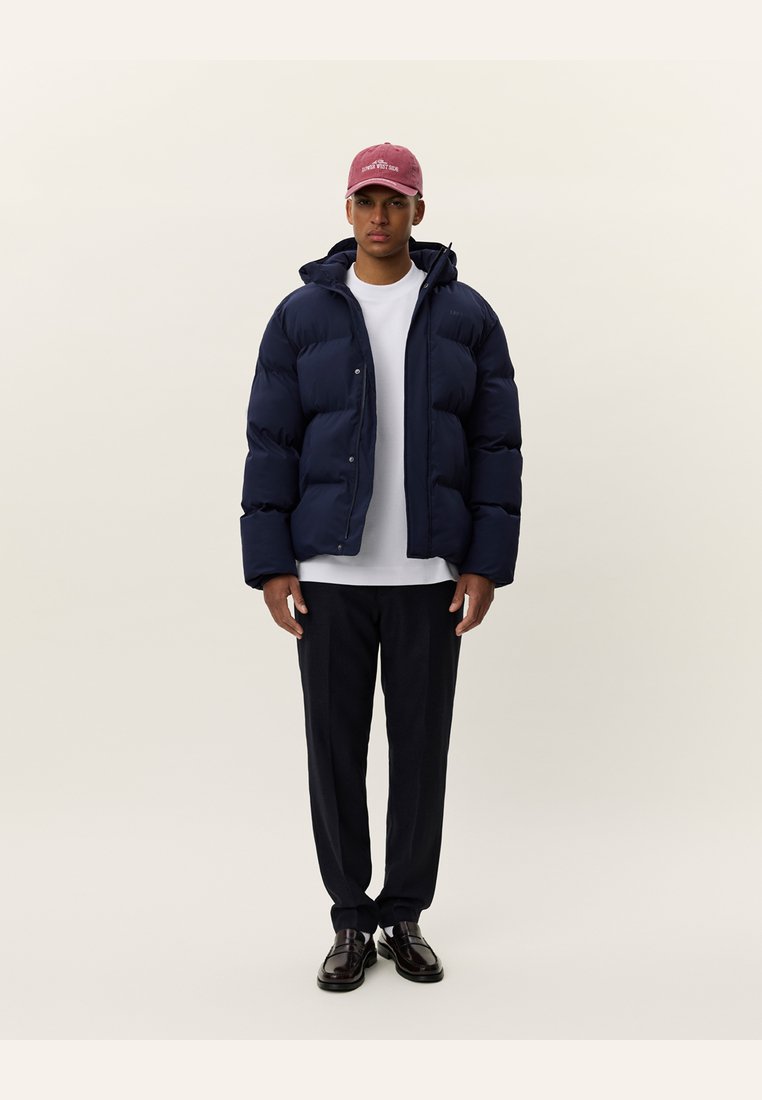 Chaqueta acolchada de color navy con diseño acolchado, capucha y botones frontales, combinada con una camiseta blanca, pantalones oscuros y zapatos negros.