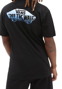 Osoba s dredy, oblečená v černém tričku Vans s modrým logem skateboardu a textem "Off The Wall" na zádech.