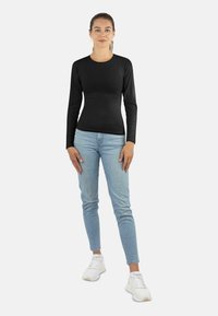 Schwarzes Langarmoberteil aus glattem Stoff, kombiniert mit hellblauen Skinny-Jeans und weißen Sneakers. Das Model steht mit entspannten Armen.