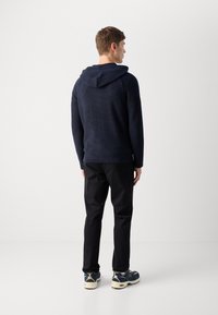 Felpa navy con un motivo testurizzato e cordoncino, abbinata a pantaloni neri e scarpe da ginnastica multicolori. Modello di spalle rispetto alla fotocamera.