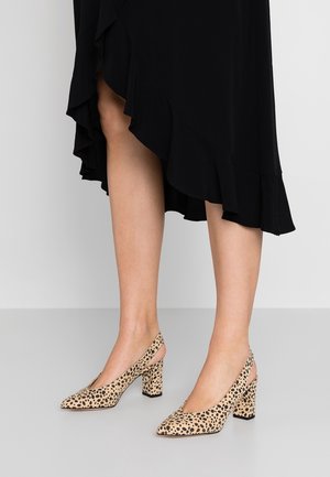 Jambes portant des talons hauts beiges à pois noirs, associés à une jupe noire à volants sur fond gris uni.