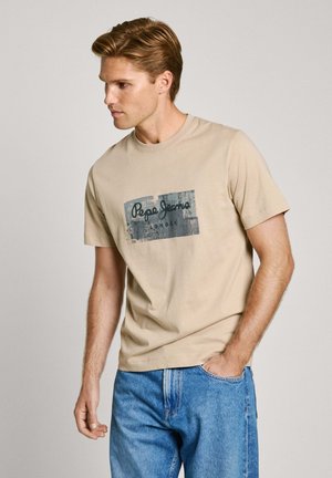Camiseta de algodón beige con un estampado gráfico que presenta "Pepe Jeans London" en azul marino. Diseño de mangas cortas y cuello redondo; combinada con unos vaqueros azules.