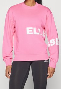 Sweat-shirt rose à manches longues et col rond. Présente le texte blanc "ELLESSE" sur le devant et la manche. À assortir avec des leggings noirs.