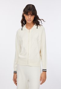 Cardigan crema in maglia con zip frontale, polsini a coste con strisce nere e motivo a rosa traforato sul corpo.