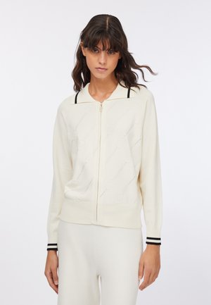 Cardigan crema in maglia con zip frontale, polsini a coste con strisce nere e motivo a rosa traforato sul corpo.