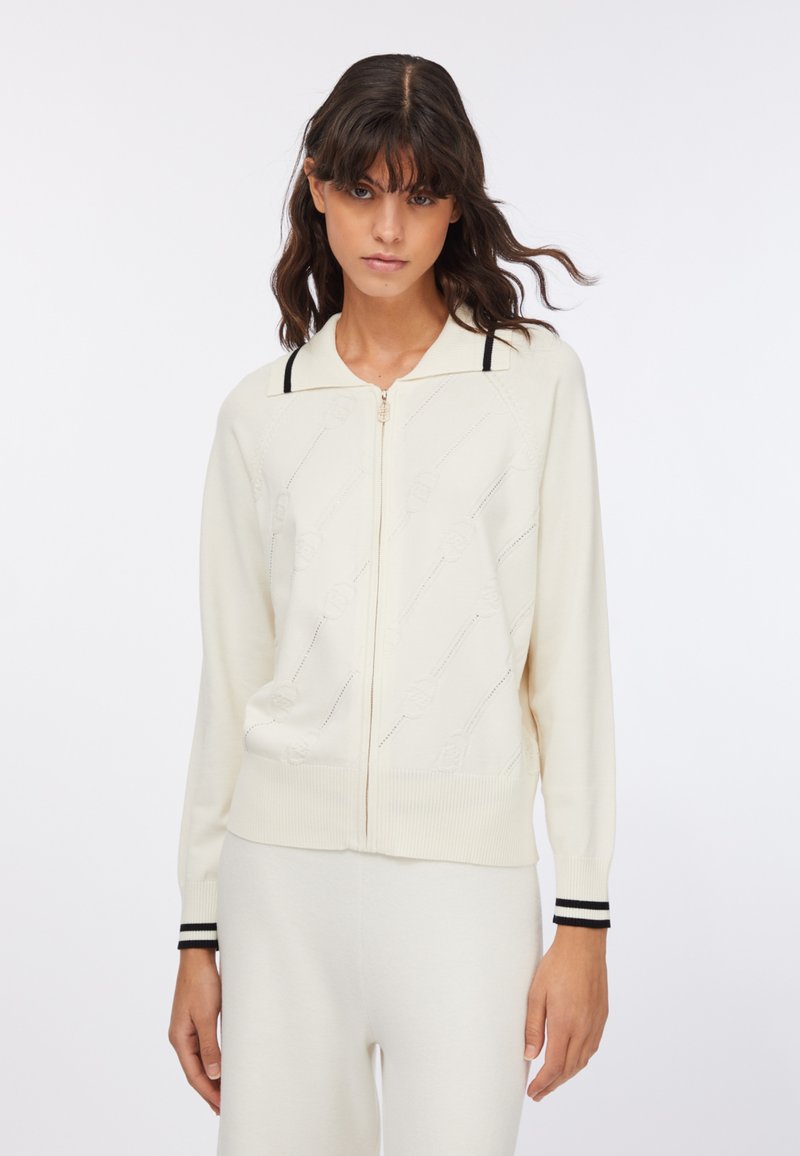 Cardigan crema in maglia con zip frontale, polsini a coste con strisce nere e motivo a rosa traforato sul corpo.