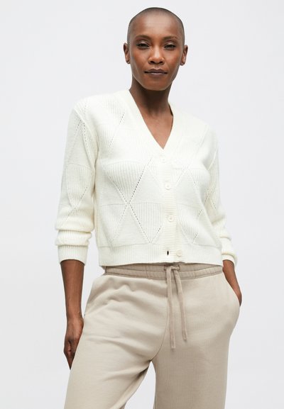 Kremowy sweter z dzianiny z dekoltem w kształcie litery V, z wzorem w romby i zapięciem na guziki, zestawiony z beżowymi spodniami dresowymi.