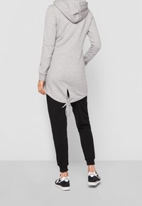 Donna che indossa una felpa lunga con cappuccio grigia chiara, dettagli con lacci sulla schiena, pantaloni jogger neri e sneakers bianco e nere della New Balance.