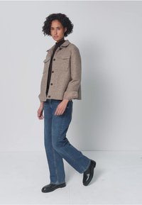 Vrouw met krullend haar draagt een beige jas met knopen over een zwart shirt, blauwe spijkerbroek en zwarte laarzen, staat met één voet iets opgetild.