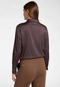 Blusa lunga a maniche, in satin marrone, con colletto classico e polsini abbottonati, abbinata a pantaloni marroni sartoriali. Texture liscia e design aderente.