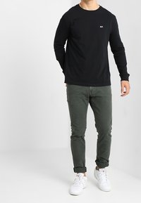 Schwarzes Langarmshirt mit Rundhalsausschnitt, kombiniert mit olivgrünen, schmal geschnittenen Hosen und weißen Sneakern. Einfaches Design mit einem kleinen Logo-Detail.