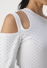 Top bianco a maniche lunghe con spalle scoperte, texture a coste e strass argentati disposti in un motivo a pois su tutta la superficie.