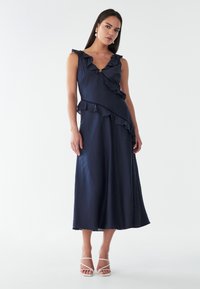 Laavupunane mekko, jossa on röyhelökoristeet kauluksessa ja helmoissa, valmistettu teksturoidusta kankaasta. Mekossa on istuva vyötärö ja virtaava hame.