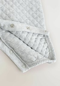 Grau gepolsterter Stoff mit einem Rautenmuster, ausgestattet mit metallischen Druckknöpfen und einem elastischen Rand, der eine glatte Textur und eine weiche Oberfläche zeigt.