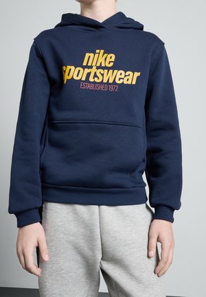 Lapsi, jolla on tummansininen Nike Sportswear -huppari keltaisella tekstillä ja vaaleanharmaat collegehousut, seisoo yksivärisen taustan edessä.