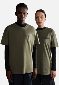 T-shirt in cotone verde oliva con colletto a coste, maniche corte e dettaglio del logo sul petto, indossato sopra maglie a maniche lunghe nere.