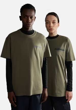 Camisetas de algodón verde oliva con cuellos redondos acanalados, mangas cortas y detalle del logo en el pecho, llevadas sobre camisetas de manga larga negras.