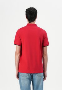 Κόκκινο πόλο μπλουζάκι με κοντό γιακά, μαύρες λεπτομέρειες στα μανίκια και μαλακή υφή. Φορεμένο με ανοιχτό γαλάζιο τζιν παντελόνι.