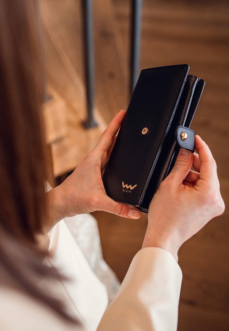 Black leather wallet held in hands, featuring a gold emblem and snap closure. Smooth texture with sleek design and subtle stitching details.