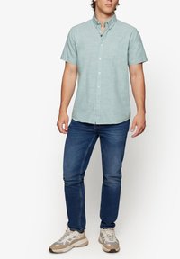 Camisa de botones de manga corta color verde claro con un bolsillo. Combinada con jeans de color azul oscuro y zapatillas de color claro con detalles en gris y beige.