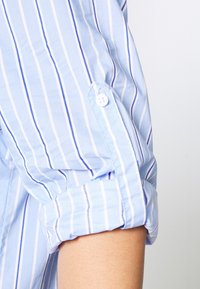Lichtblauw button-up shirt met witte en donkerblauwe verticale strepen, met een opgerolde mouw en een kleine knop op de manchette.
