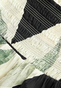 Gros plan sur un tissu avec un smockage élastique froncé, présentant des motifs abstraits noirs, crème et verts, ainsi que des lignes de couture parallèles blanches.